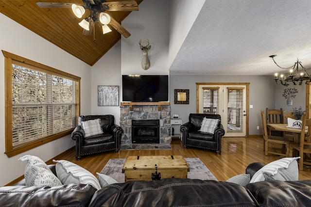 living room featuring high vaulted ceiling, a stone fireplace, ceiling fan with notable chandelier, wood finished floors, and baseboards