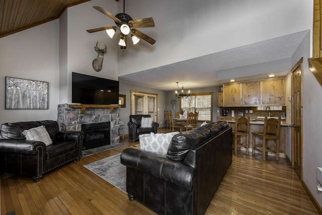 living room featuring high vaulted ceiling, wood-type flooring, ceiling fan with notable chandelier, and a stone fireplace