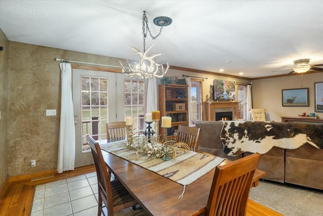 dining room featuring a warm lit fireplace, light tile patterned floors, baseboards, crown molding, and ceiling fan with notable chandelier