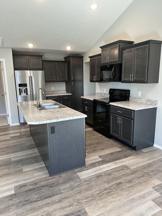 kitchen featuring lofted ceiling, wood finished floors, a sink, black appliances, and a center island with sink