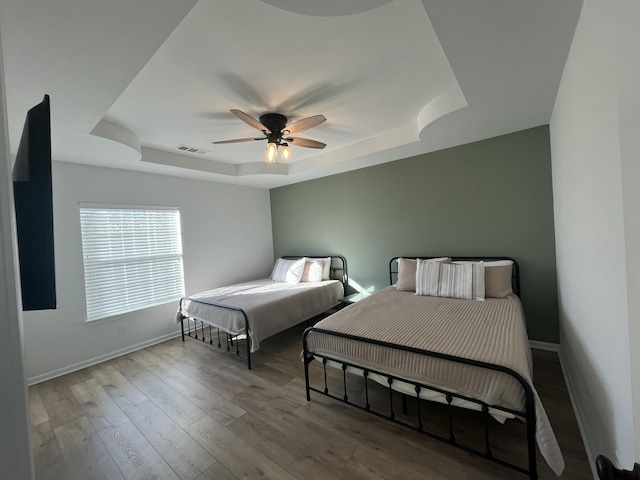 bedroom with a ceiling fan, wood finished floors, visible vents, baseboards, and a tray ceiling