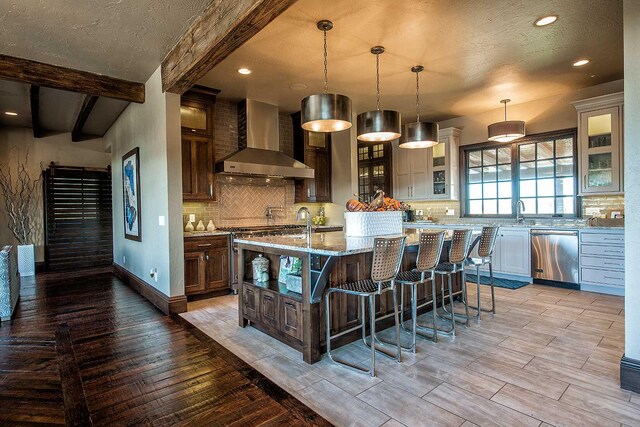 kitchen with stainless steel dishwasher, wood tiled floor, wall chimney exhaust hood, beamed ceiling, and glass insert cabinets