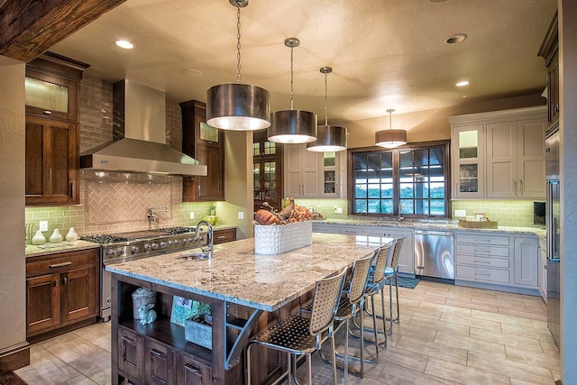 kitchen with high end appliances, backsplash, an island with sink, light stone countertops, and wall chimney exhaust hood