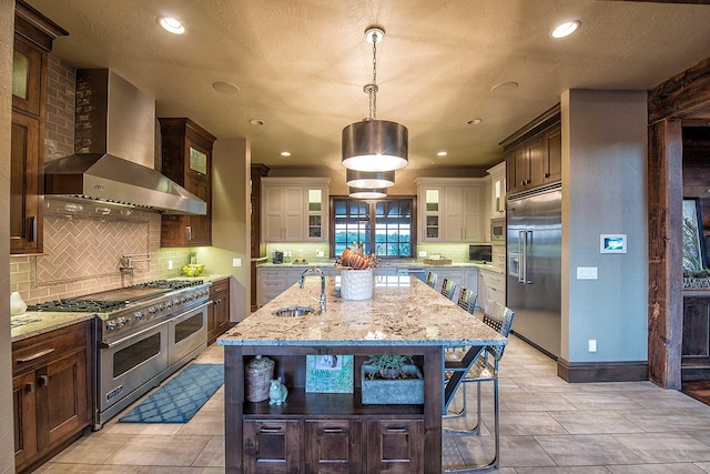 kitchen with a kitchen island with sink, a sink, wall chimney range hood, light stone countertops, and built in appliances