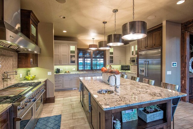 kitchen featuring a kitchen island with sink, a sink, wall chimney range hood, and built in appliances