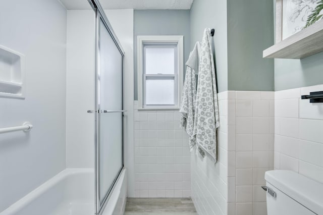 bathroom featuring toilet, a wainscoted wall, bath / shower combo with glass door, wood finished floors, and tile walls