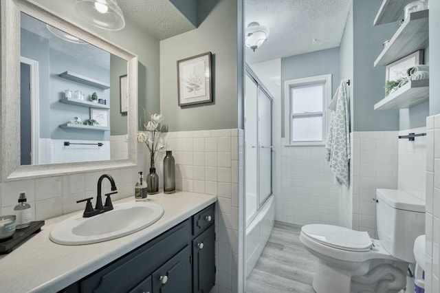 bathroom with tile walls, toilet, wainscoting, combined bath / shower with glass door, and a textured ceiling