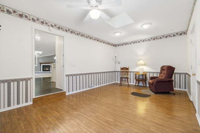 sitting room featuring a skylight, wood finished floors, a ceiling fan, baseboards, and a lit fireplace