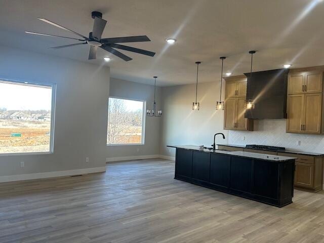 kitchen with baseboards, light wood-style floors, range hood, decorative backsplash, and a center island with sink