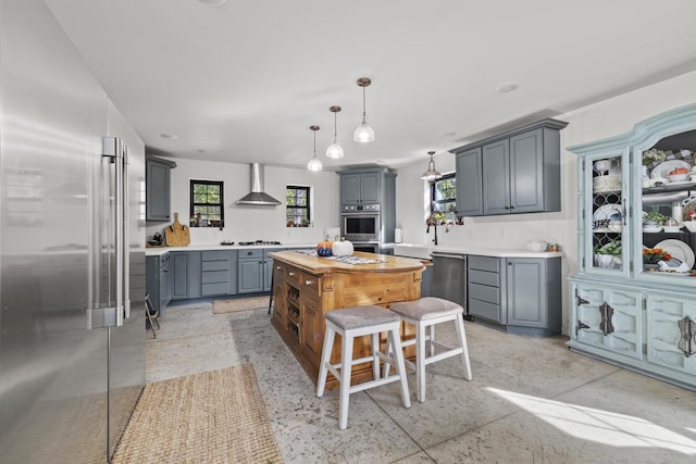 kitchen featuring gray cabinetry, stainless steel appliances, hanging light fixtures, a center island, and wall chimney exhaust hood