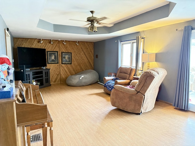 living area featuring wooden walls, a raised ceiling, a ceiling fan, an accent wall, and wood finished floors