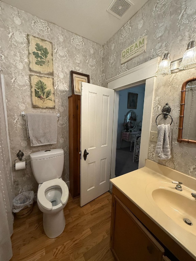 bathroom featuring visible vents, toilet, vanity, wood finished floors, and wallpapered walls