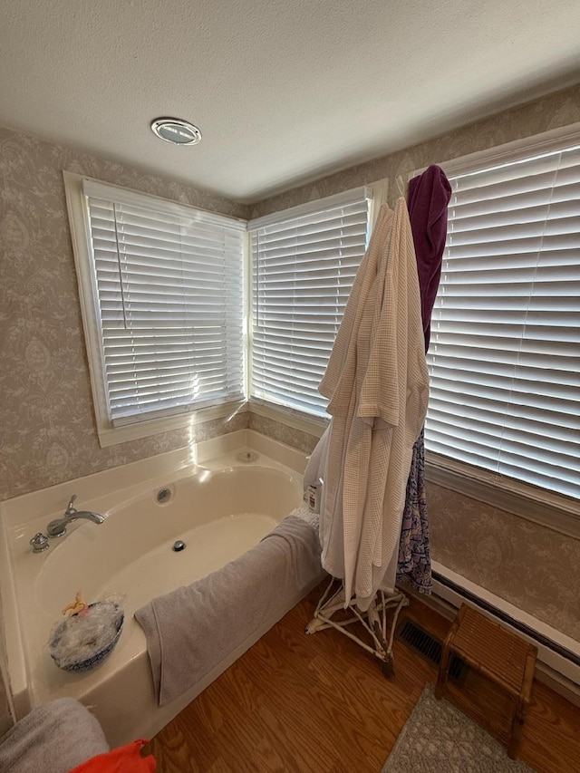 bathroom with wallpapered walls, baseboard heating, a textured ceiling, wood finished floors, and a bath