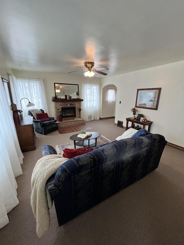 living room with baseboards, arched walkways, a ceiling fan, a fireplace with raised hearth, and carpet flooring