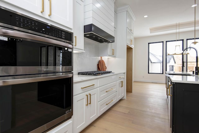 kitchen featuring light wood-style flooring, appliances with stainless steel finishes, white cabinets, a sink, and wall chimney exhaust hood