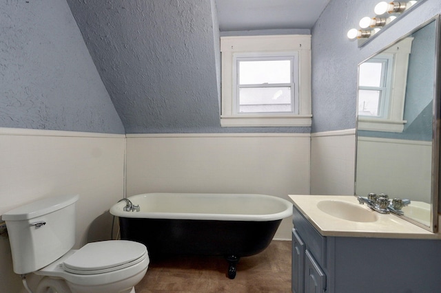 bathroom with a textured wall, toilet, vanity, a freestanding bath, and wainscoting