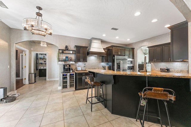 kitchen featuring beverage cooler, arched walkways, custom range hood, light stone counters, and stainless steel appliances