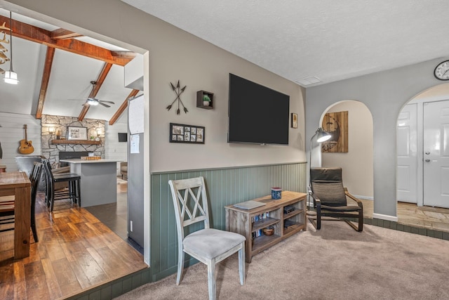 living area with arched walkways, a ceiling fan, beamed ceiling, a textured ceiling, and carpet flooring