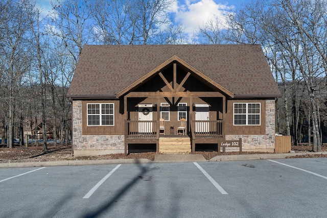 chalet / cabin with a porch, stone siding, roof with shingles, and uncovered parking