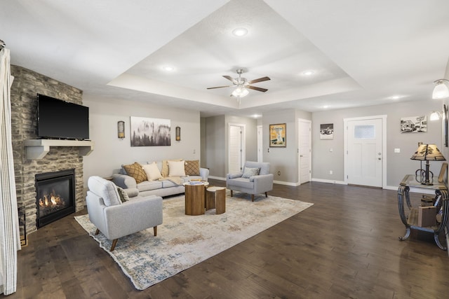 living area with baseboards, ceiling fan, dark wood-style flooring, a tray ceiling, and a fireplace