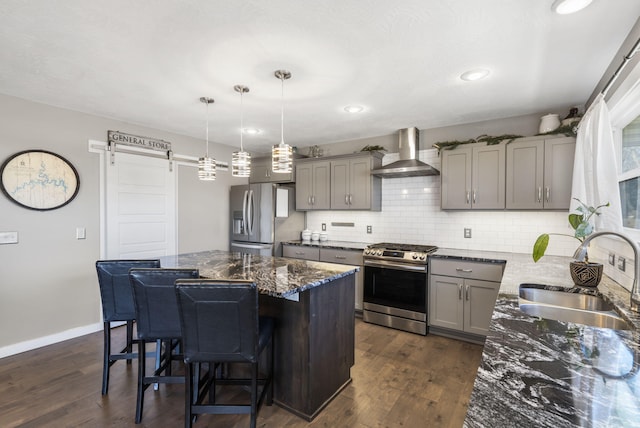 kitchen with a barn door, gray cabinetry, a sink, appliances with stainless steel finishes, and wall chimney exhaust hood