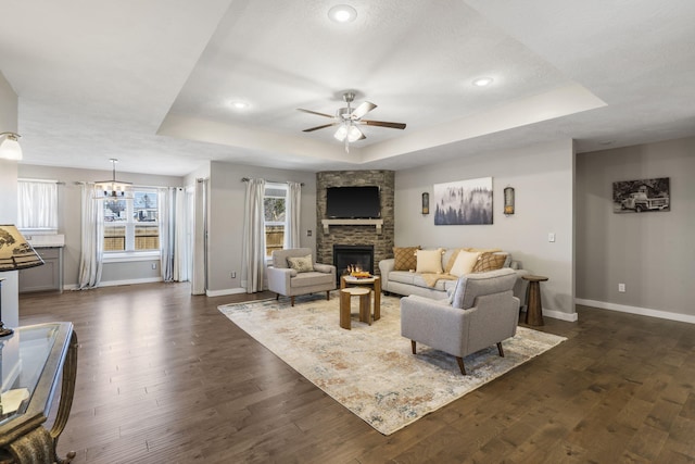 living room featuring a stone fireplace, a tray ceiling, dark wood-style flooring, and a ceiling fan