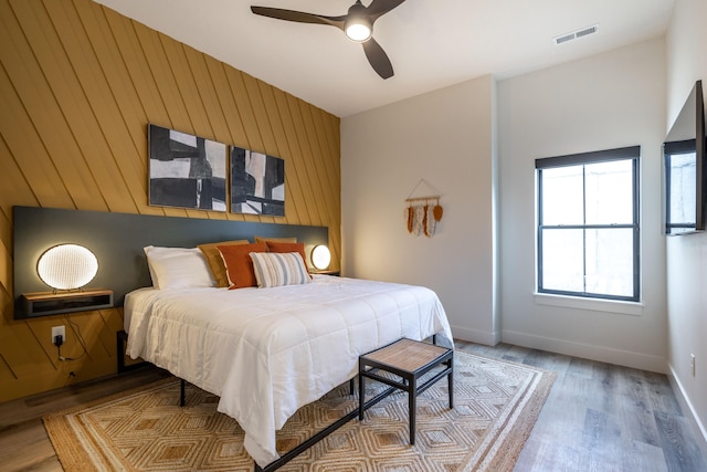 bedroom with ceiling fan, wood walls, visible vents, baseboards, and light wood-type flooring