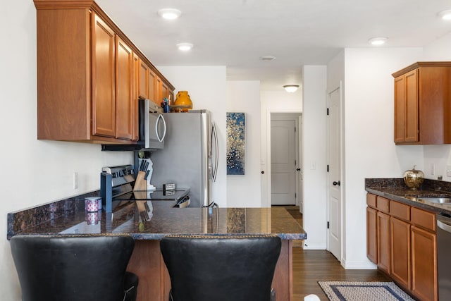 kitchen featuring dark wood-type flooring, stainless steel appliances, dark stone counters, a peninsula, and brown cabinetry