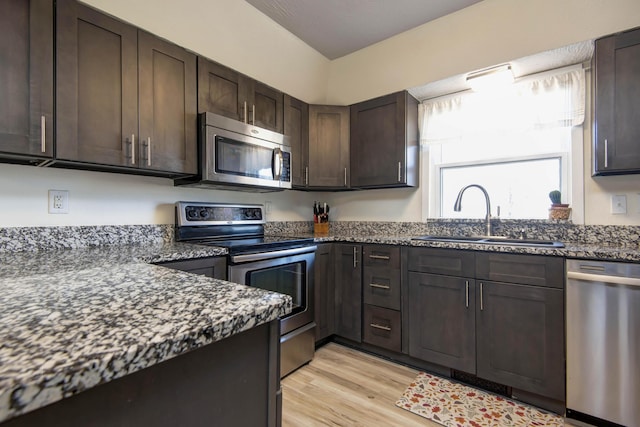 kitchen with light wood-style flooring, a sink, dark brown cabinets, appliances with stainless steel finishes, and dark stone counters