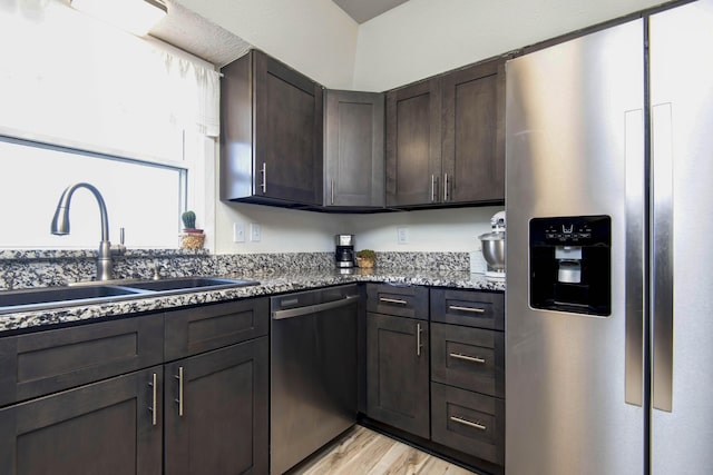 kitchen with light wood-style flooring, appliances with stainless steel finishes, a sink, dark brown cabinetry, and dark stone counters