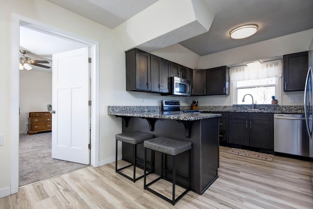 kitchen with light wood finished floors, stainless steel appliances, a sink, dark stone counters, and a kitchen bar