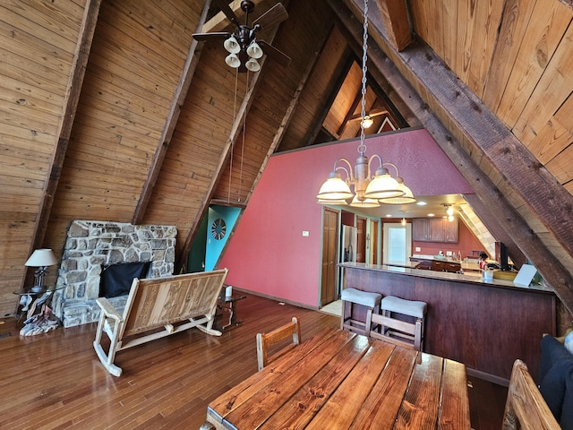 dining area featuring a stone fireplace, ceiling fan with notable chandelier, wood finished floors, wood ceiling, and beamed ceiling