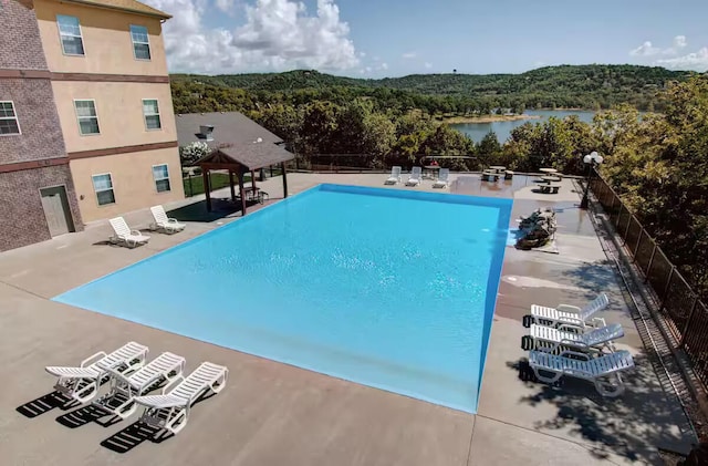 community pool with a patio area, a water view, fence, and a view of trees