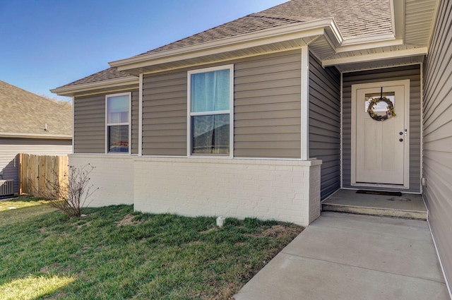 doorway to property with brick siding, a shingled roof, fence, and a yard