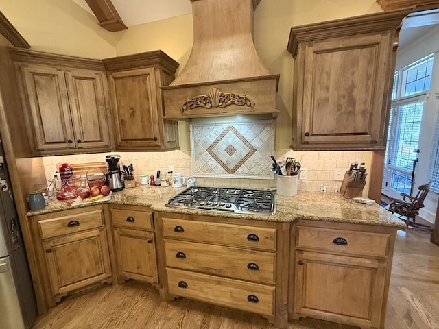 kitchen with light stone counters, premium range hood, light wood-style flooring, stainless steel gas stovetop, and tasteful backsplash