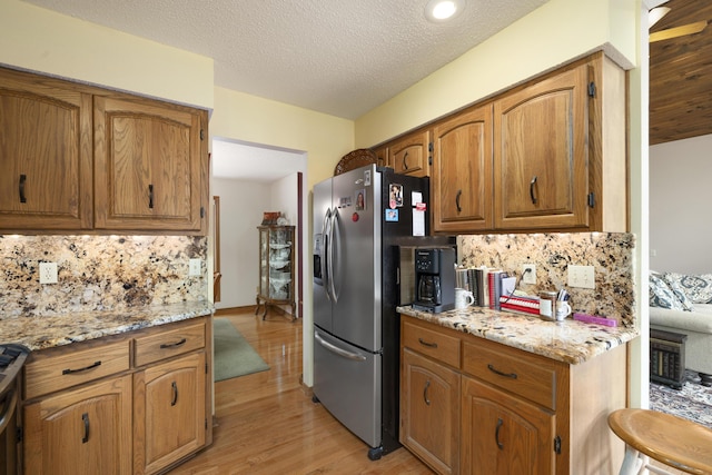 kitchen featuring brown cabinets, stainless steel refrigerator with ice dispenser, backsplash, light stone countertops, and light wood-type flooring