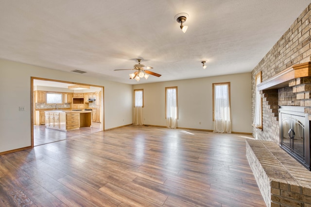 unfurnished living room featuring a brick fireplace, wood finished floors, visible vents, and a ceiling fan