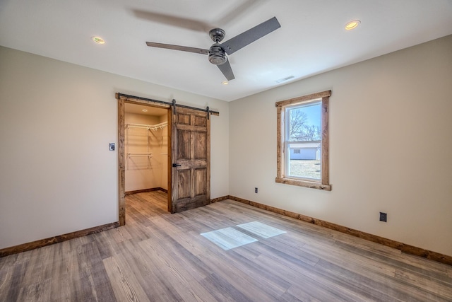 unfurnished bedroom featuring a barn door, a ceiling fan, baseboards, a spacious closet, and light wood-type flooring