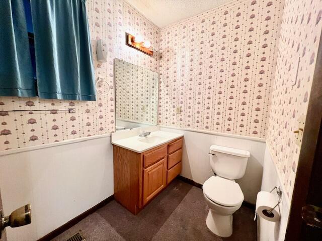 bathroom with toilet, a textured ceiling, wainscoting, wallpapered walls, and vanity