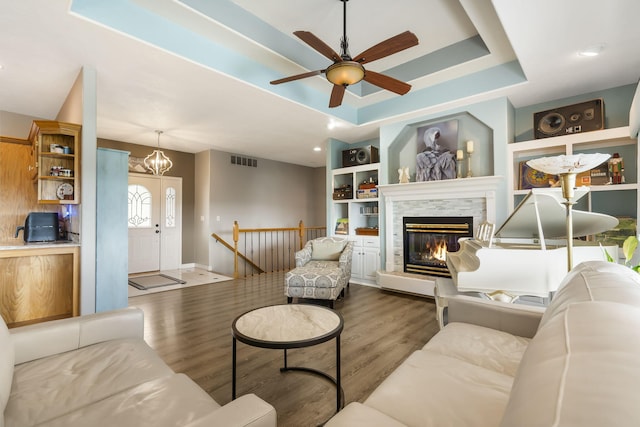 living room featuring built in features, wood finished floors, visible vents, a glass covered fireplace, and a raised ceiling