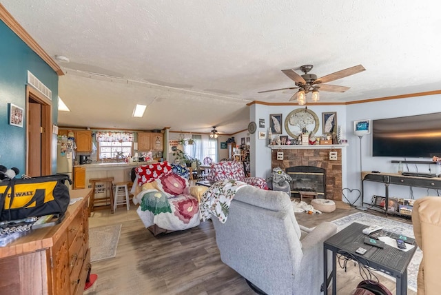 living room featuring a brick fireplace, light wood-type flooring, ornamental molding, a textured ceiling, and a ceiling fan