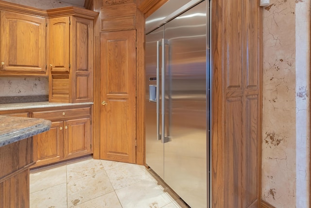 kitchen with light tile patterned floors, stainless steel built in fridge, light stone countertops, and brown cabinetry