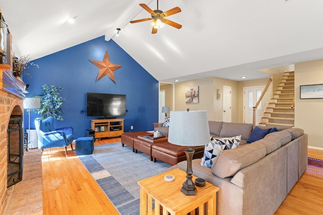 living area with light wood finished floors, a brick fireplace, ceiling fan, beamed ceiling, and stairway