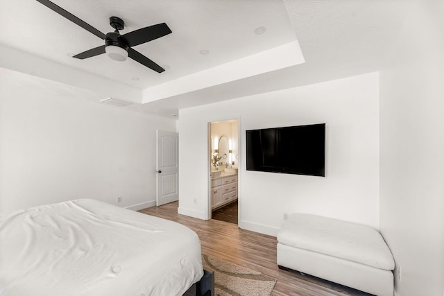 bedroom featuring a tray ceiling, ensuite bath, wood finished floors, baseboards, and ceiling fan