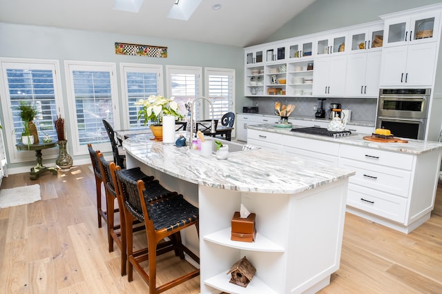 kitchen with a center island with sink, open shelves, light wood-style flooring, a sink, and appliances with stainless steel finishes