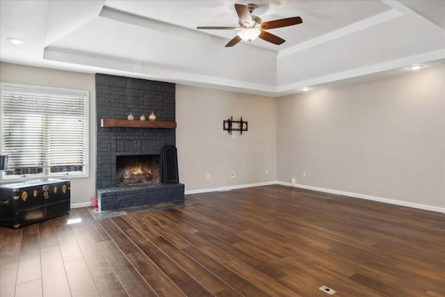 unfurnished living room featuring wood finished floors, baseboards, ceiling fan, a raised ceiling, and a brick fireplace