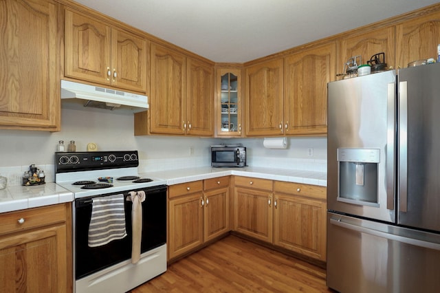 kitchen featuring under cabinet range hood, appliances with stainless steel finishes, brown cabinetry, glass insert cabinets, and tile counters