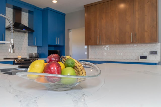 kitchen featuring blue cabinets, decorative backsplash, light stone countertops, and wall chimney range hood