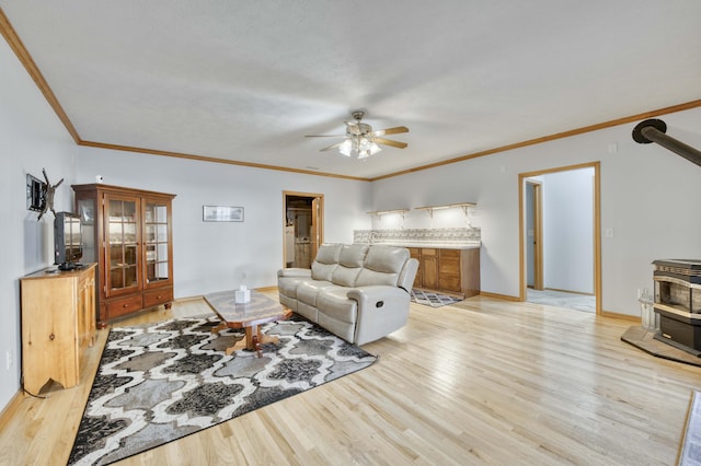 living room featuring light wood-type flooring, ornamental molding, a ceiling fan, baseboards, and a wood stove