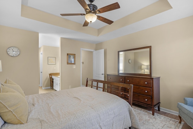 bedroom with baseboards, light wood finished floors, a tray ceiling, ceiling fan, and connected bathroom